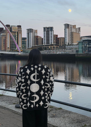 Person wearing a patterned coat standing by a waterfront with a city skyline in the background.