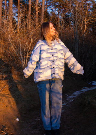 Person wearing a white cloud embroidered jacket cardigan standing in a forest with trees and blue sky.